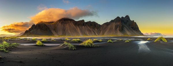 Im Vordergrund erstreckt sich ein schwarzer Sandstrand, hinter dem sich im Hintergrund ein schroffes Bergmassiv erhabt.