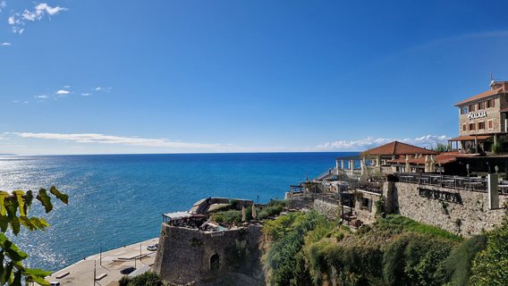 Von einem Hügel, der rechts mit einem Restaurant mit Außenterrasse aus großen Steinquadern bebaut ist, blickt man weit über das leuchtendblaue Meer. Darüber erstreckt sich ein fast wolkenloser strahlendblauer Himmel.