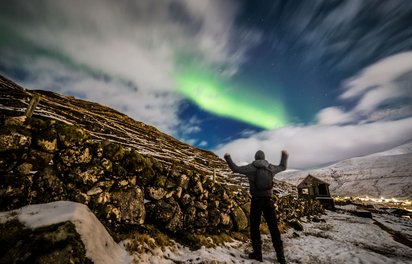 Das Nordlicht begeistert Etwa mittig steht ein Mann mit erhobenen Armen; man sieht ihn in Rückenansicht, während er ein grünes Nordlicht im Bildhintergrund betrachtet.
