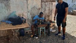 An einer Straßenecke sitzt ein Mann mit einer Nähmaschine und leistet Schneiderarbeiten.