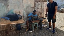 An einer Straßenecke sitzt ein Mann mit einer Nähmaschine und leistet Schneiderarbeiten.