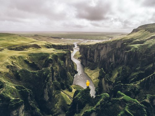 Vogelperspektive auf einen von grün bewachsenen Steilwänden eingeschlossenen Canyon, vorne in der Mitte steht klein ein Mensch in einer gelben Jacke.