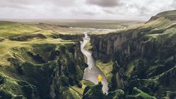 Vogelperspektive auf einen von grün bewachsenen Steilwänden eingeschlossenen Canyon, vorne in der Mitte steht klein ein Mensch in einer gelben Jacke.