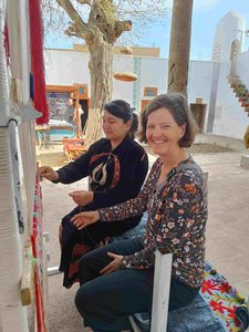 An einem Webstuhl aug einem Platz zeigt eine alte Usbekin einer in die Kamera lachenden Touristin ihr Handwerk.