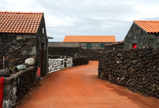 Ein rot-brauner von Mauern und Häusern aus schwarzem Lavagestein gesäumter Weg führt in den Bildhintergrund.