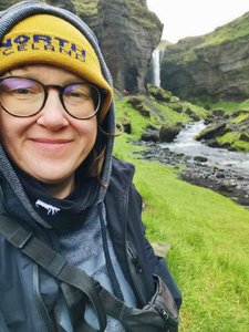 Selbstporträt vor dem Kvernufoss-Wasserfall in Island Eine lächelnde Person mit Brille und gelber Mütze steht vor einer grünen Schlucht. Im Hintergrund stürzt der Kvernufoss-Wasserfall über eine hohe Felswand in ein Bachbett.