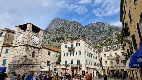 Um einen kleinen Platz stehen mehrere große, historische Gebäude mit Fassaden aus großen, grauen Quadersteinen oder gelb verputzt. Dahinter erhebt sich ein Berg und darüber blauer Himmel mit Schleierwolken.