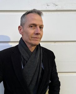 Portrait eines Mannes mit grauen Haaren, schwarzer Jacke und schwarzem Schal.