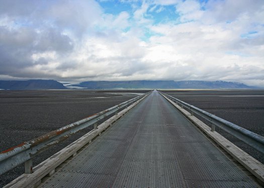 Unter einem wolkenverhangenen, aber blauen Himmel zieht sich ein Teil der isländischen Ringstraße hin, das sich in der Ferne vor einer Hügellandschaft verliert.