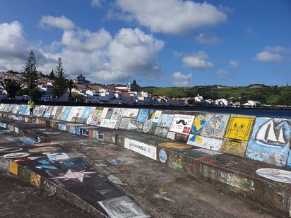 Im Vordergrund durchzieht eine bunt bemalte Kaimauer das Foto, im Hintergrund erheben sich die Häuser von Horta.