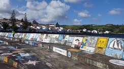 Im Vordergrund durchzieht eine bunt bemalte Kaimauer das Foto, im Hintergrund erheben sich die Häuser von Horta.