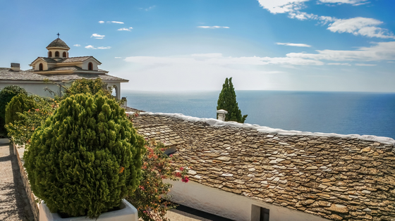 Links im Vordergrund stehen Büsche, dahinter sieht man ein kirchenartiges Gebäude in weiß mit grauen Schieferdächern. Die rechte vordere Bildhälfte wird ebenfalls von einem Schieferdach dominiert, hinter dem sich der Blick auf das blaue Meer öffnet-