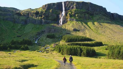 Wanderung zum Bjarnarfoss-Wasserfall auf Snæfellsnes Zwei Personen wandern auf einem Wiesenpfad in Richtung eines hohen Wasserfalls, der sich über steile Klippen und Basaltsäulen hinabstürzt. Im Vordergrund grünes Gras, im Hintergrund Felsen und Wald.