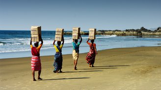 Frauen in traditioneller Kleidung am Strand tragen Kisten auf ihren Köpfen 
