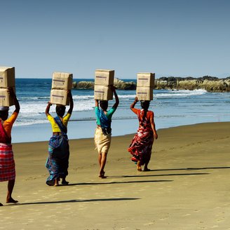 Frauen in traditioneller Kleidung am Strand tragen Kisten auf ihren Köpfen 