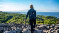 Eine vom Betrachter abgewendete Frau steht auf einem felsigen Untergrund und schaut auf das im Hintergrund befindliche Meer.