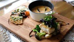 Auf einem Holzbrett ist eine Schale mit Fischsuppe sowie ein einzelnes Stück Fisch mit Gemüse und Salat arrangiert.