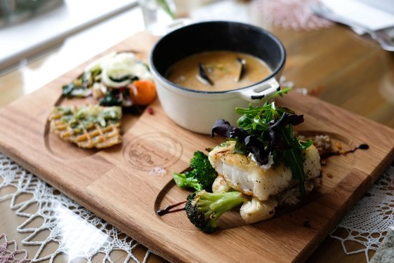 Auf einem Holzbrett ist eine Schale mit Fischsuppe sowie ein einzelnes Stück Fisch mit Gemüse und Salat arrangiert.