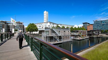 Hausboote im Viertel Västra Hamnen Auf einem Kanal liegen drei schwimmende Häuser aus Holz, dahinter befindet sich moderne Bebauung.