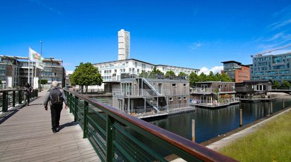 Auf einem Kanal liegen drei schwimmende Häuser aus Holz, dahinter befindet sich moderne Bebauung.