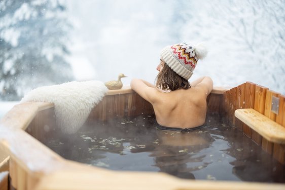 Eine Frau mit einer Pudelmütze sitzt in einem Badezuber vor einer verschneiten Landschaft.
