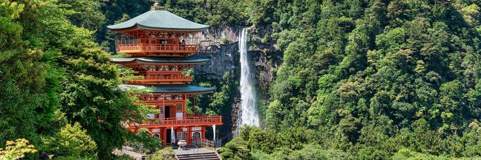 Blick auf einen dreistöckig roten Tempel, der inmitten eines dichten, grünen Waldes steht. Im Hintergrund befindet sich ein Wasserfall an einer Steinmauer.
