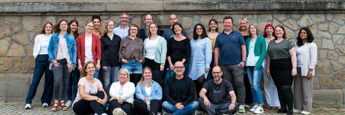 Das Reisen Mit Sinnen Team und contrastravel Team steht und sitzt gemeinsam vor einer Mauer und schaut frontal in die Kamera.