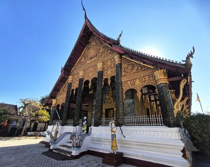 Tempel in Luang Prabang DAs Bild wird fast komplett von einem spitzgiebeligen Tempel ausgefüllt. Auf einem Fundament aus weißem Stein erheben sich verzierte Säulen mit goldenen Kapitellen, die den Giebel stützen. Der gesamte Tempel ist üppig goldverziert. Darüber erhebt sich ein leuchtend blauer Himmel.
