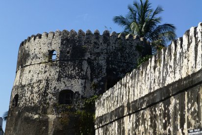Festung in Stone Town Blick entlang einer Mauer auf einen Festungsturm, dahinter eine Palmkrone und blauer Himmel.