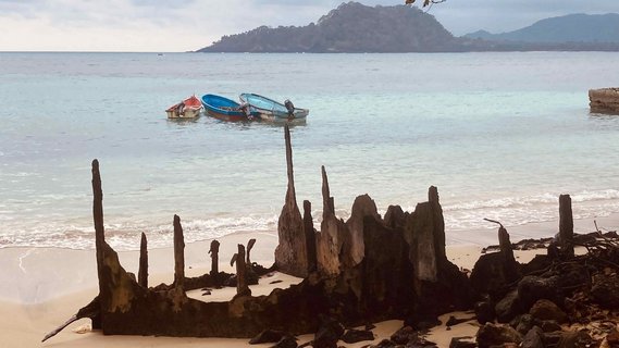 Strand von São Tomé und Príncipe Heller Strand, an dem rostige Streben eines Transportschiffes aus dem Sand ragen. Im Hintergrund ist das blaue Meer mit kleinen Booten und die Umrisse einer Insel.