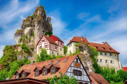 Im vorderen Bild sieht man den Giebel eines Fachwerkhauses, dahinter staffelt sich ein burgartiges Gebäude an einem Berghang, der mit einer Felsnadel vor blauem Himmel endet.