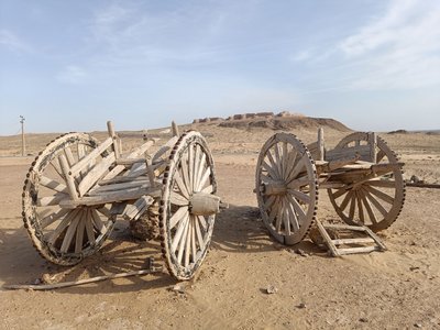 An der Wüstenfestung Ayaz Kala Im Vordergrund sieht man zwei Karren, die von einem Lasttier gezogen werden können, im Hintergrund am Horizont erhebt sich Ton in Ton mit dem Sand eine Festung. Etwa das obere Drittel des Bildes besteht aus blauem Himmel.