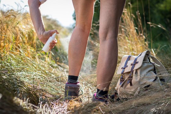 Eine Frau sprayt ihre in Wanderschuhen steckenden, aber sonst unbekleideten Beine mit Mückenspray ein.