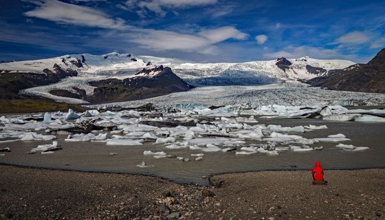Vorne rechts sitzt ein Mensch in einer roten Wetterjacke auf einem Stein und schaut auf einen Gletschersee, hinter dem sich der Gletscher ertreckt.