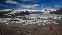 Vorne rechts sitzt ein Mensch in einer roten Wetterjacke auf einem Stein und schaut auf einen Gletschersee, hinter dem sich der Gletscher ertreckt.