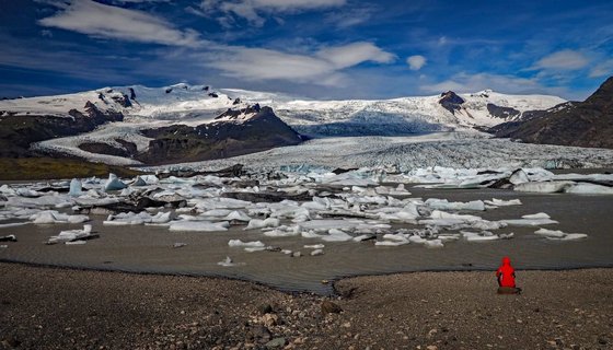 Vorne rechts sitzt ein Mensch in einer roten Wetterjacke auf einem Stein und schaut auf einen Gletschersee, hinter dem sich der Gletscher ertreckt.