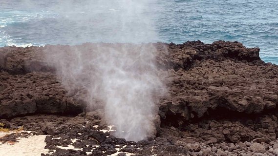 Dunkle Felsen aus denen der Qualm der heißen Quellen hoch steigt. Im Hintergrund ist das blaue Meer.