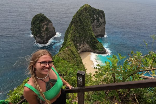 Ann-Kathrin Schmitz steht auf einer Treppe. Im Hintergrund ist die grüne Inselküste und das blaue Meer von Nusa Penida.