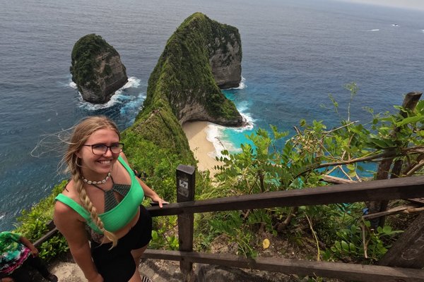 Ann-Kathrin Schmitz steht auf einer Treppe. Im Hintergrund ist die grüne Inselküste und das blaue Meer von Nusa Penida.