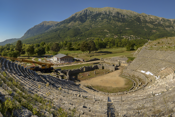 Das Amphitheater von Dodona Aus den oberen Rängen eines großen Amphitheaters schaut man hinunter auf die Bühne, hinter der sich eine grüne, baumbestanden Landschaft erstreckt. Dahinter erhebt sich eine grüne Bergkette, darüber blauer Himmel.