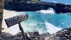 Seitlicher Blick auf eine Bucht, in der der weiße Sand von links nach rechts in die Wellen des blauen Meeres verläuft. Im Vordergrund steht ein Schild mir der Beschriftung "Praia Escada".