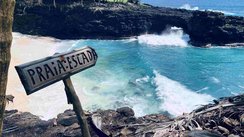 Seitlicher Blick auf eine Bucht, in der der weiße Sand von links nach rechts in die Wellen des blauen Meeres verläuft. Im Vordergrund steht ein Schild mir der Beschriftung "Praia Escada".