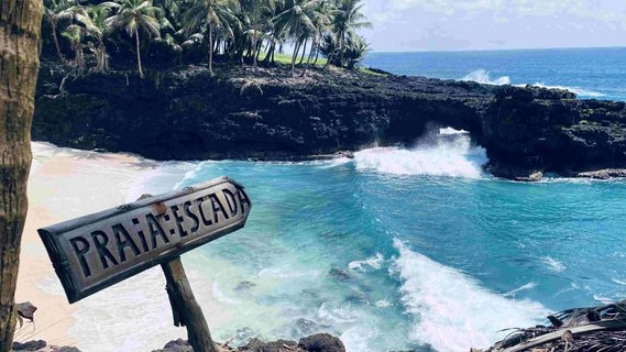 Seitlicher Blick auf eine Bucht, in der der weiße Sand von links nach rechts in die Wellen des blauen Meeres verläuft. Im Vordergrund steht ein Schild mir der Beschriftung "Praia Escada".