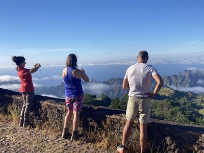 Drei Wanderer sehen mit dem Rücken zum Betrachter vor einer gut kniehohen Mauer und schauen hinab in das dahinterliegende grüne Tal. Die Frau ganz links macht ein Foto von der Aussicht vom Cova Corda über Santo Antao. Die obere Bildhälfte wird von blauem Himmel eingenommen.