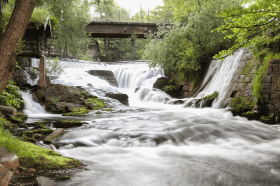 Über ein Wehr stürzt ein Wasserlauf, fließt auf den Betrachter zu und nimmt den gesamten vorderen Bildraum ein. Im Hintergrund ist er von Bäumen umfasst und von einer Brücke überspannt.