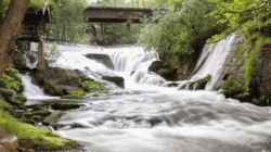 Über ein Wehr stürzt ein Wasserlauf, fließt auf den Betrachter zu und nimmt den gesamten vorderen Bildraum ein. Im Hintergrund ist er von Bäumen umfasst und von einer Brücke überspannt.