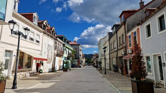 Blick in eine Straßenflucht, die von niedrigen, maximal zweigeschossigen Häusern flankiert ist. In einigen Häusern befinden sich im Erdgeschoss Geschäfte mit großen Schaufenstern. Der Himmel darüber ist blau mit großen weiß-grauen Wolken.
