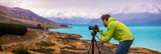 Auf Fotoreisen die Welt entdecken Vor einer Berg- und Wasserlandschaft steht ein Mann in einer grünen Jacke und schaut auf den Monitor einer Kamera, die auf einem Stativ befestigt ist.