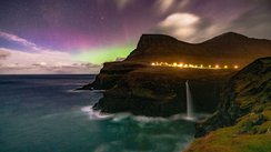 Unter einem lila und grün leuchtendem Himmel erhebt sich eine Klippe mit einem Wasserfall aus dem Meer.