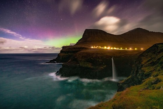 Nordlichter über Vagar Unter einem lila und grün leuchtendem Himmel erhebt sich eine Klippe mit einem Wasserfall aus dem Meer.